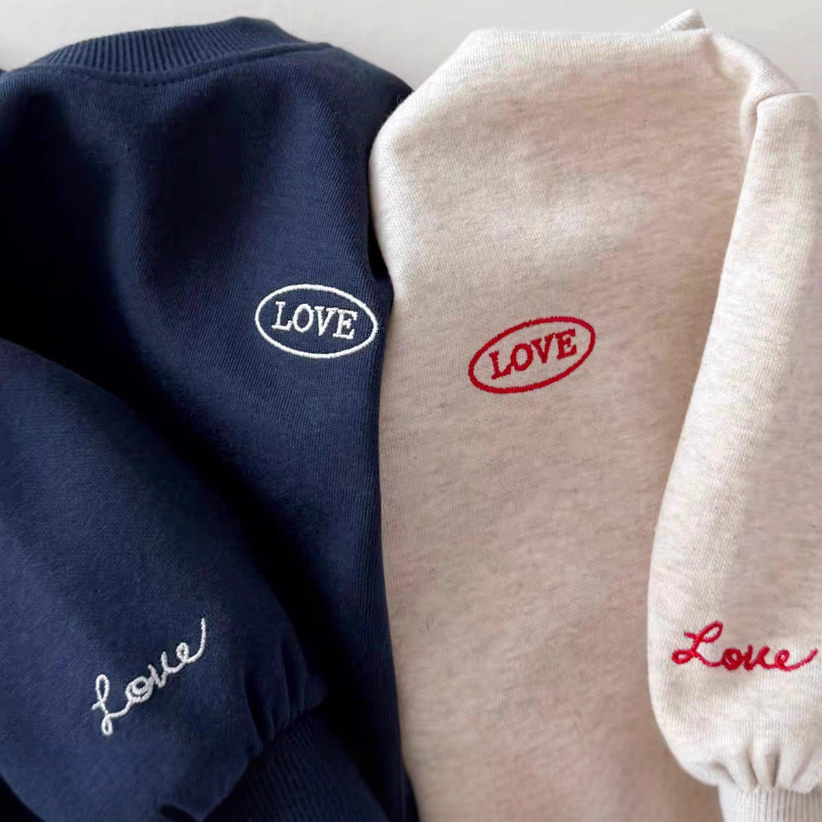 Two sweatshirts, one navy and one beige, with 'LOVE' embroidery on a light gray background.