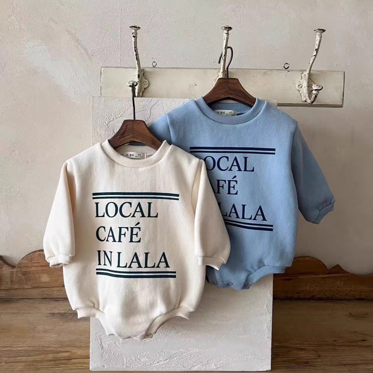 Two baby rompers, one white and one blue, with text on hangers against a neutral wall.