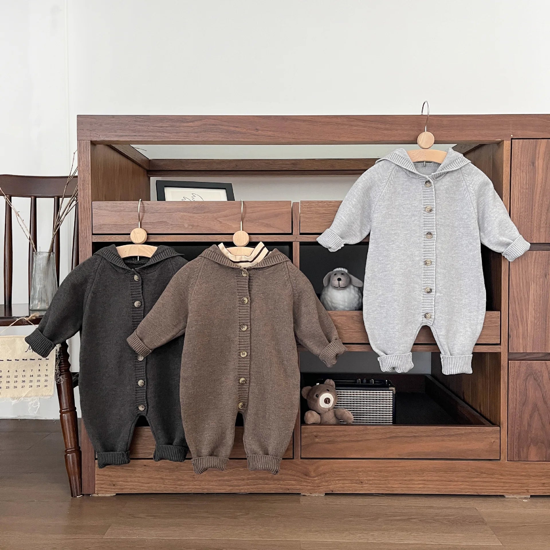 Three baby rompers hanging on a wooden wardrobe with a teddy bear inside.